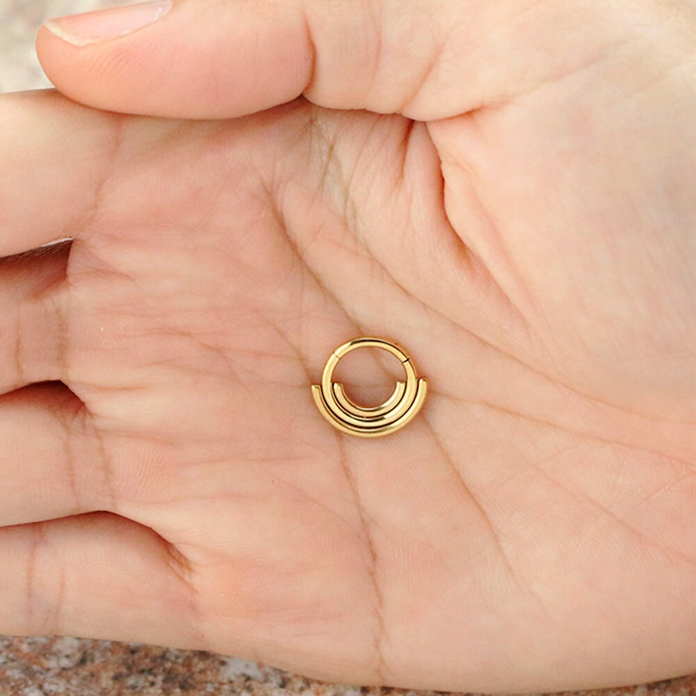 A close-up image of a hand holding the Lira Minimalist Boho Chic Septum in 316L Steel Gold PVD Finish from Kaya Movement. The palm is open, showcasing the refined gold Bohemian jewelry piece with its modern style. The background is blurred and indistinct, highlighting the septum's elegant design and craftsmanship.
